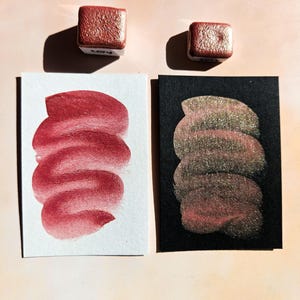 Puede incluir: Dos pastillas de acuarela cuadradas y muestras de color. Las pastillas de pintura son de color marrón rojizo metálico. Las muestras de pintura están sobre papel blanco y negro. La muestra de papel blanco es de color marrón rojizo sólido, y la muestra de papel negro tiene un brillo dorado.