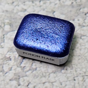 May include: A square, deep blue watercolour paint pan with a shimmering, textured surface. The paint is in a white plastic container with the words "POTION FLASK" printed on the front.