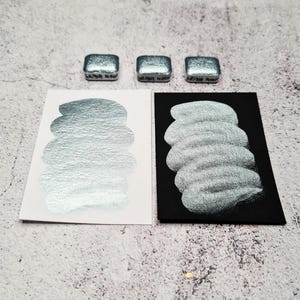 Puede incluir: Tres pastillas de pintura de acuarela cuadradas, de color verde azulado metálico, se muestran sobre muestras del mismo color sobre papel blanco y negro. Las muestras muestran la calidad brillante y nacarada de la pintura. La pintura está etiquetada como "Bright Moon".