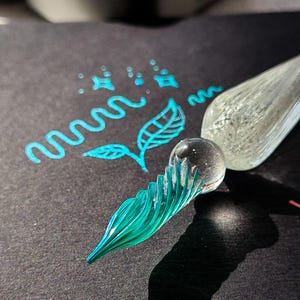 May include: A clear glass dip pen with a teal-colored nib, casting a shadow on a dark surface. Teal ink drawings of a leaf, wavy lines, and stars are next to the pen. A small metal paint palette is in the background.