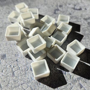 Może przedstawiać: Kolekcja małych, białych, kwadratowych palet do malowania. Każda paleta ma płytką, otwartą komorę. Palety są rozrzucone na teksturowanej, szarej powierzchni, z rzucanymi cieniami.