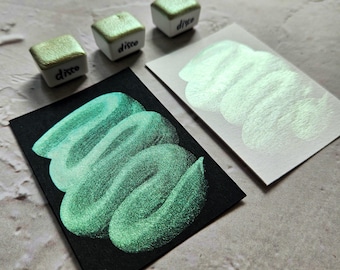 May include: An overhead shot showcases three small, square paint pans labeled "disco" with a shimmering green hue. Swatches of the same color are displayed on black and white cards, demonstrating the paint's iridescent quality. The paint appears to have a metallic finish.