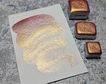 Puede incluir: Una muestra de pintura de acuarela brillante en tonos dorados y rosas sobre una tarjeta blanca. También se ven tres pequeños godets de pintura cuadrados con el texto "CUTIE PIE", que muestran el mismo color metálico.