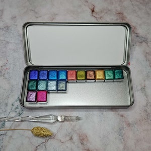 Könnte beinhalten: Eine silberne Metall-Farbpalette mit 20 Aquarellfarben. Die Farben sind in einer Reihe angeordnet und enthalten eine Vielzahl von Farben, darunter Blau, Grün, Gold und Pink. Die Palette ist geöffnet und die Farben sind sichtbar. Ein Glas-Tintenfüller liegt auf einer Marmorfläche neben der Palette.