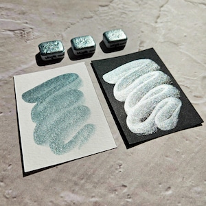 May include: Three small, square watercolour paint pans with teal metallic paint are displayed above two swatches. One swatch is on white paper, the other on black paper. The swatches show the paint's colour and shimmer.