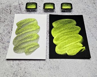 Puede incluir: Tres pastillas de pintura de acuarela cuadradas etiquetadas como "Teal Mess" se muestran encima de dos muestras. Una muestra está sobre papel blanco, la otra sobre papel negro. Ambas muestras muestran una pincelada verde brillante y metálica.