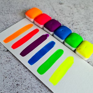Op de afbeelding: Een set van acht neon waterverfverven in een regenboog van kleuren, waaronder oranje, rood, paars, blauw, groen en geel. De verven zitten in kleine, vierkante verpakkingen en zijn in een rij op een wit oppervlak gerangschikt.