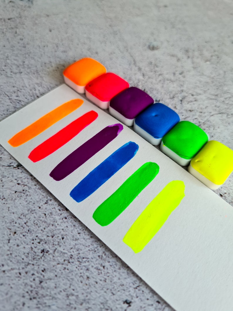 Op de afbeelding: Een set van acht neon waterverfverven in een regenboog van kleuren, waaronder oranje, rood, paars, blauw, groen en geel. De verven zitten in kleine, vierkante verpakkingen en zijn in een rij op een wit oppervlak gerangschikt.