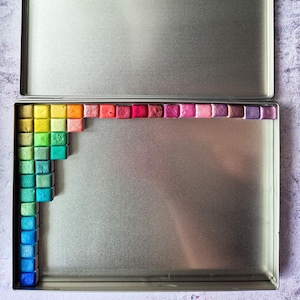 May include: A rectangular silver metal tin containing a collection of small, square watercolor pans arranged in an L-shaped border along the top and left edges. The pans display a gradient of colors ranging from yellow and orange to pink, purple, blue, and green.