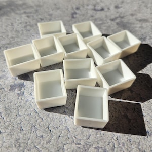 Könnte beinhalten: Elf kleine, cremeweiße, rechteckige Farbpasten sind auf einer strukturierten, grauen Oberfläche verstreut. Jede Palette hat eine quadratische Form mit einem vertieften Innenraum. Schatten verleihen der Anordnung Tiefe.