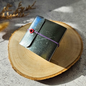 May include: A small, square, metallic-looking box with a flap closure secured by a purple elastic band and a red grommet. The box is resting on a round wooden slice, with a neutral background.