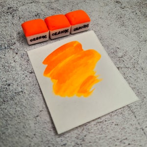 Op de afbeelding: Drie kleine oranje verfpotjes met het woord "Oranje" erop geschreven. Een staal van feloranje verf op een wit vel papier.