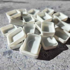 Può includere: Una collezione di piccoli contenitori di plastica bianchi e rettangolari. Ogni contenitore ha una forma quadrata leggermente arrotondata con un interno incassato. I contenitori sono sparsi su una superficie grigia strutturata.