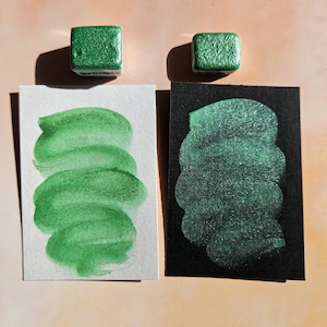 Puede incluir: Dos pastillas de pintura de acuarela verde metálico cuadradas se muestran sobre muestras en papel blanco y negro. Las muestras muestran el color y la textura de la pintura. La pintura parece tener un efecto brillante.