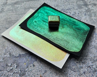 May include: A close-up of watercolor paint samples. The image shows a small, square, metallic green paint cube on a black card with a vibrant green paint swatch. A white card with a gold paint swatch is below.