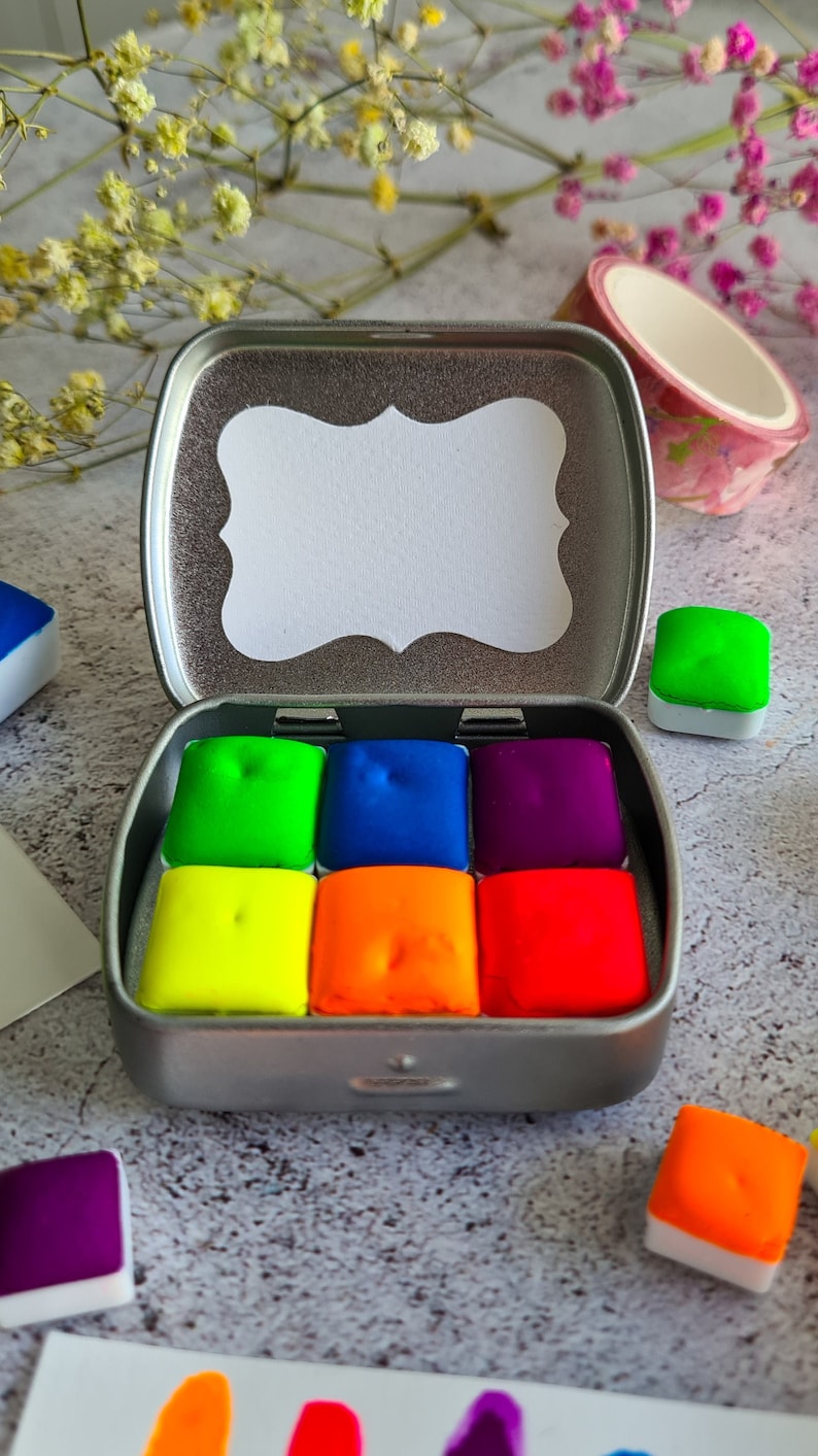 Op de afbeelding: Een metalen doosje met een wit label en een regenboog van aquarelverfpannetjes. De pannetjes zijn gerangschikt in een 3x2 raster en zijn in de kleuren groen, blauw, paars, geel, oranje en rood.