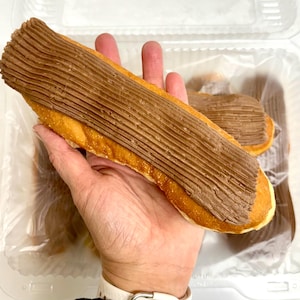 May include: A hand holding a long, golden-brown pastry topped with a thick layer of chocolate frosting. Another similar pastry is visible in the background, inside a clear plastic container. The frosting has a ridged texture.