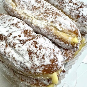 May include: Close-up of several long, golden-brown pastries dusted with powdered sugar. The pastries have a creamy filling visible at the ends. They are arranged in a clear plastic container, suggesting a bakery or dessert shop.