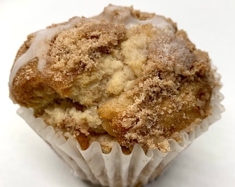 Könnte beinhalten: Nahaufnahme eines Zimt-Streusel-Muffins mit weißem Zuckerguss. Der Muffin befindet sich in einer weißen Papierform und liegt auf einer weißen Oberfläche.