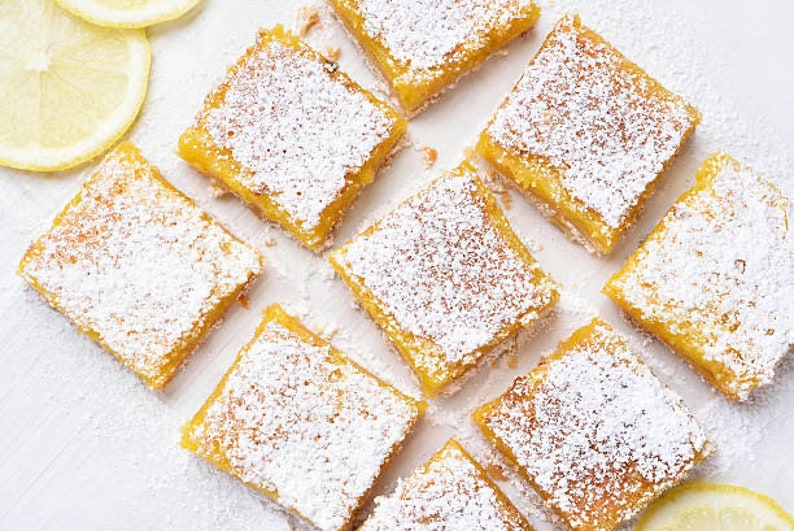 Peut inclure: Gros plan sur neuf barres au citron, chacune saupoudr&eacute;e de sucre glace, dispos&eacute;es sur une surface blanche. Les barres sont carr&eacute;es et de couleur jaune clair.