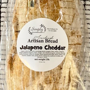 May include: A loaf of Jalapeno Cheddar Artisan Bread from Simply Sourdough Co. in a clear plastic bag. The bread is a light brown color and has a slightly crispy crust. The label on the bag reads "Simply Sourdough Co. Artisan Bread Jalapeno Cheddar" and lists the ingredients and allergens. The bread weighs 2 pounds.