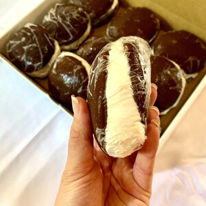 May include: A close-up of a chocolate-covered marshmallow treat with a white filling. The treat is wrapped in plastic and held in a hand. The treat is one of many in a box.