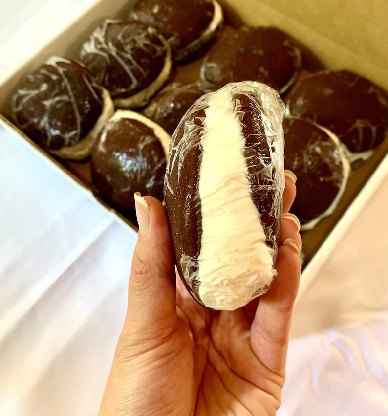 May include: A close-up of a chocolate-covered marshmallow treat with a white filling. The treat is wrapped in plastic and held in a hand. The treat is one of many in a box.