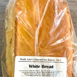 May include: A white loaf of gluten-free white bread in a clear plastic bag. The label reads "Ruth Ann's Gluten-Free Bakery, LLC" and "White Bread". The ingredients and nutritional information are listed on the label. The label also includes the address "5620 Umbletown Rd - Gap, PA 17527 (PA Dept. of Ag.)".