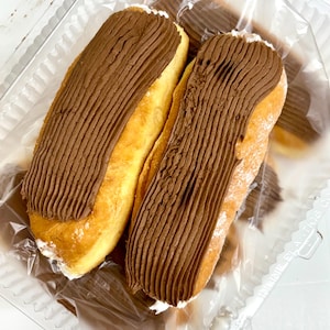 May include: Two oblong pastries with golden-brown exteriors and a thick layer of chocolate frosting. The frosting is textured with parallel lines. The pastries are in a clear plastic container, suggesting they are ready to eat.