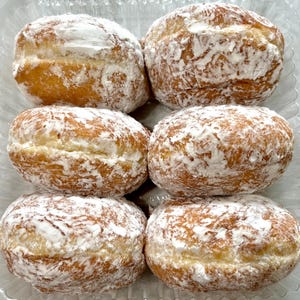 May include: Six powdered sugar donuts in a clear plastic container. The donuts are golden brown with a generous coating of white powdered sugar. The donuts are round and appear freshly made.