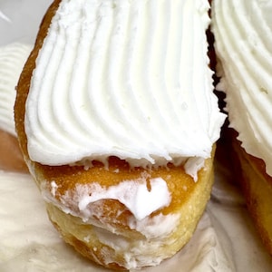 May include: Close-up of a cream-filled pastry. The pastry has a golden-brown exterior and is topped with a generous swirl of white whipped cream. The cream appears fluffy and textured, contrasting with the smooth pastry.