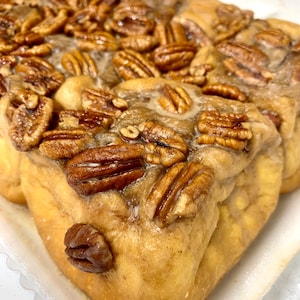 May include: Close-up of a pecan sticky bun with a golden brown crust and a generous amount of pecans on top. The bun is glazed with a sweet, sticky glaze.