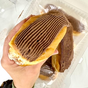 May include: A hand holding a golden-brown pastry with a thick layer of chocolate frosting. The pastry is in a clear plastic container, with a second pastry visible in the background. The frosting is a dark brown color and has a ridged texture.