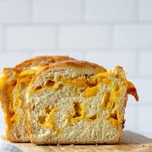 May include: Two slices of golden-brown cheese bread on a wooden cutting board. The bread has a soft, white interior with pockets of melted orange cheese. The crust is lightly toasted, and the bread is ready to eat.