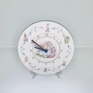 Puede incluir: Un plato de cerámica blanco con un diseño floral y una esfera de reloj con una ilustración de un gato y un conejo. La esfera del reloj tiene números romanos y manecillas marrones.