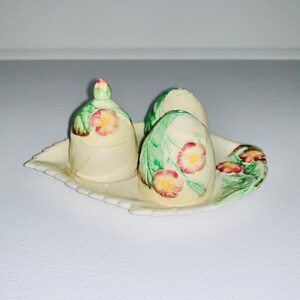 Könnte beinhalten: Ein Set aus drei Salz- und Pfefferstreuern aus Keramik mit floralem Design. Die Streuer sind weiß mit grünen Blättern und rosa Blüten. Sie stehen auf einem passenden weißen Tablett mit gewelltem Rand.