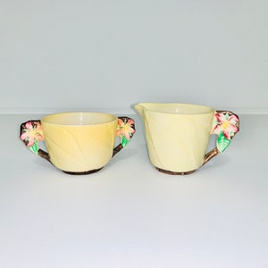 Könnte beinhalten: Ein Vintage-Sahne- und Zuckerset. Die Teile sind hellgelb mit blattartigen Texturen. Jeder Griff ist ein brauner Ast mit rosa und weißen Blüten und grünen Blättern. Das Set steht auf einer weißen Oberfläche.