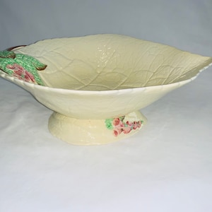 Puede incluir: Cuenco de cerámica color crema con forma de hoja y base de pedestal. El cuenco tiene una superficie texturizada con detalles de venas de hoja. La base está decorada con detalles florales rosas y verdes. El borde de la hoja tiene un acento marrón.