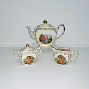 Könnte beinhalten: Ein weißes Porzellan-Tee-Set mit goldenen Akzenten. Die Teekanne, die Milchkännchen und die Zuckerdose haben ein florales Design und ein zentrales Bild eines Paares in einem Garten.