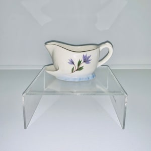 May include: A white ceramic gravy boat with a blue and purple floral design. The boat is sitting on a clear acrylic stand.