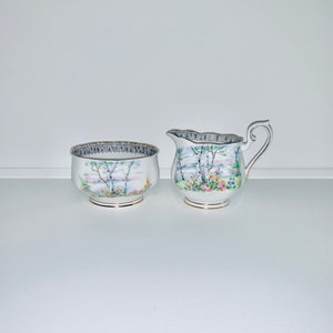 Puede incluir: Un juego de azucarero y jarra de porcelana vintage. El juego presenta un diseño de paisaje con árboles, flores y un cuerpo de agua. Los bordes están adornados con un patrón gris y plateado, y las bases tienen detalles dorados.