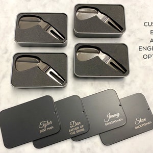 May include: Four black metal boxes with engraved names, including "Tyler Best Man", "Dan Father of the Bride", "Jimmy Groomman", and "Steve Groomman". Each box contains a silver golf divot tool. The boxes are arranged in two rows of two. The text "CUSTOM BOX AND ENGRAVING OPTIONS" is written in white on a gray background.