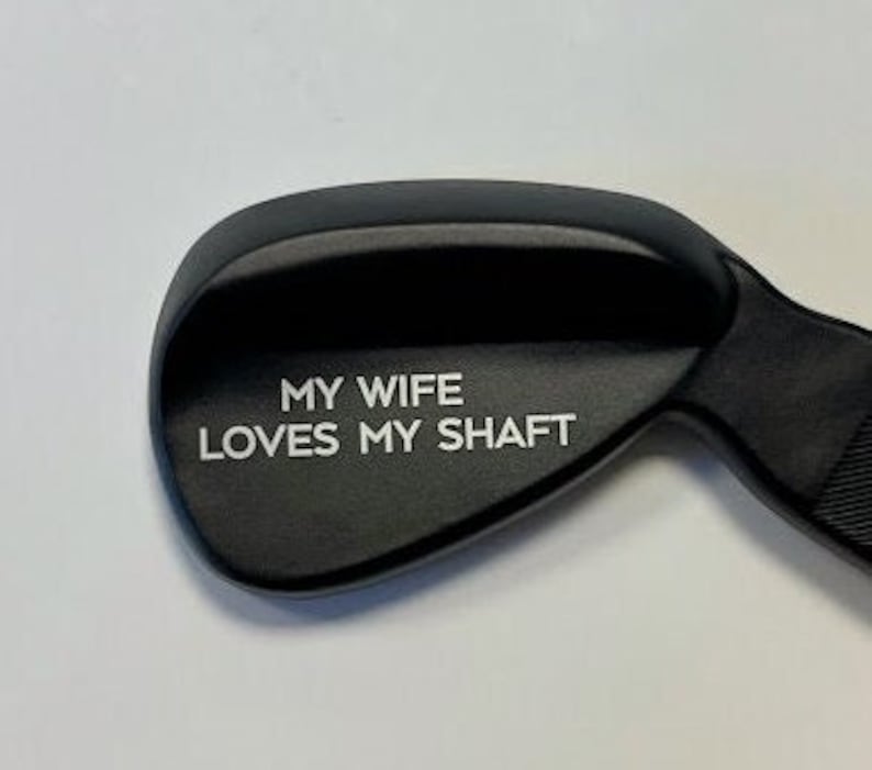 May include: A black golf club wedge with the text "My wife loves my shaft" engraved on the face.