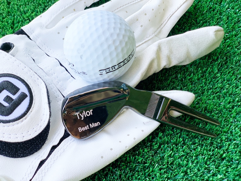 May include: A black golf ball marker with the text "Tyler Best Man" engraved on it. The marker is lying on a white golf glove with a golf ball.