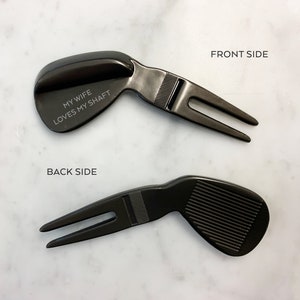 May include: Black golf ball divot tool with a golf club head design. The back side of the tool has the text "My wife loves my shaft".