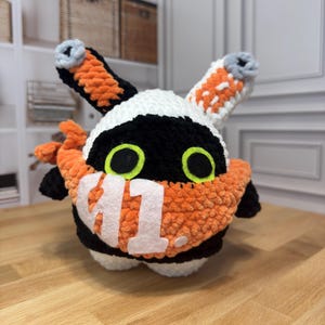 May include: A handmade, plush toy in the shape of a creature with orange, black, and white crochet. The toy has large, green eyes and the number "41" in white on its chest. The ears are orange and black with gray tips.