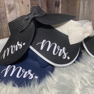 May include: Three straw sun hats with a wide brim and a bow on the top. The hats are black, white, and navy blue. Each hat has the word "Mrs." written on the front in white cursive lettering.