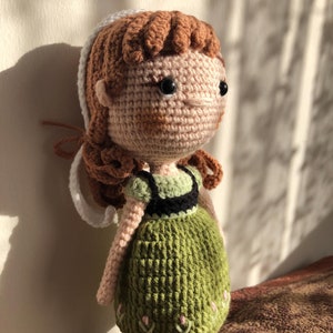 May include: Crocheted doll with brown hair, a white headband, and a green dress with floral embroidery. The doll has black eyes and a pink nose.