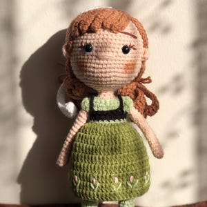 May include: A crocheted doll with brown hair, a green dress, and black shoes. The doll has a white crocheted hat and a smiling face.