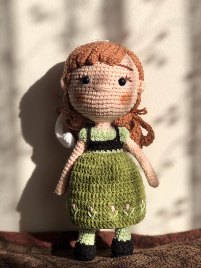 May include: A crocheted doll with brown hair, a green dress, and black shoes. The doll has a white crocheted hat and a smiling face.